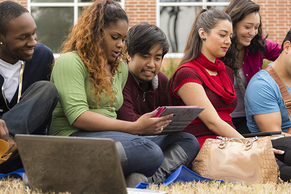 Multi-ethnic group of college student friends. Digital tablet, laptop. Multi-ethnic group of college student friends hang out on campus. They are using digital tablets and laptop computers as they sit on grass in front of a school building. The group of university friends are enjoying the fall afternoon visiting together.