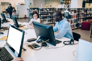 Attentive young multiethnic man and woman in casual outfit talking and doing homework while sitting together at table with computer with empty screen in library